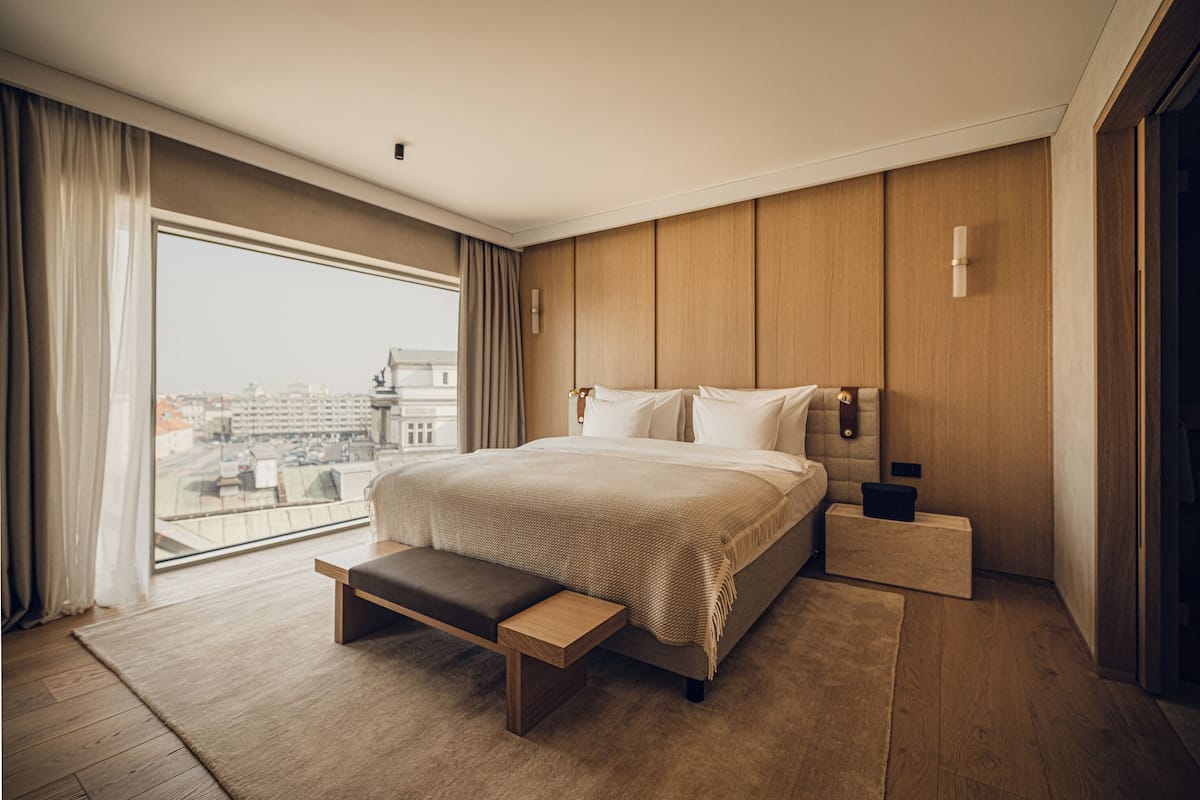 Ein modernes Hotelzimmer mit einem großen Bett, einer Bank, einem Nachttisch und Blick auf die Stadt.