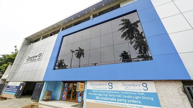 Un bâtiment polyvalent doté d’une grande façade en verre réfléchissant, d’une enseigne indiquant le « Restaurant-café Signature 9 » et d’un panneau publicitaire annonçant différents espaces événementiels, tels qu’une salle de conférence et un petit espace dédié aux cérémonies de mariage.