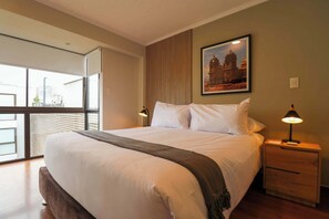 A hotel room with a large bed, a nightstand with a lamp, a window with a view of a cityscape, and a framed picture on the wall. - w * | Magnificent 3BR in the Heart of Barranco (Barranco)