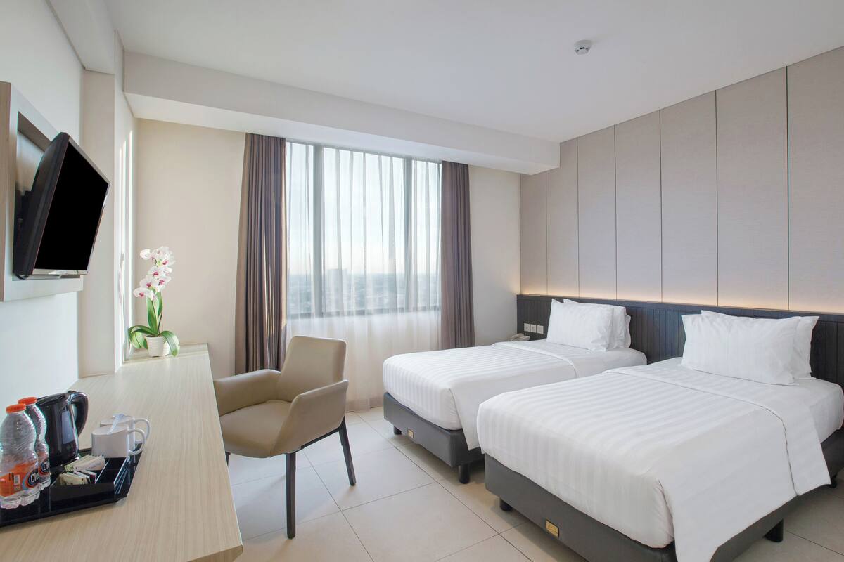 Habitación de hotel con dos camas, un escritorio con silla, televisión y una ventana grande con cortinas.