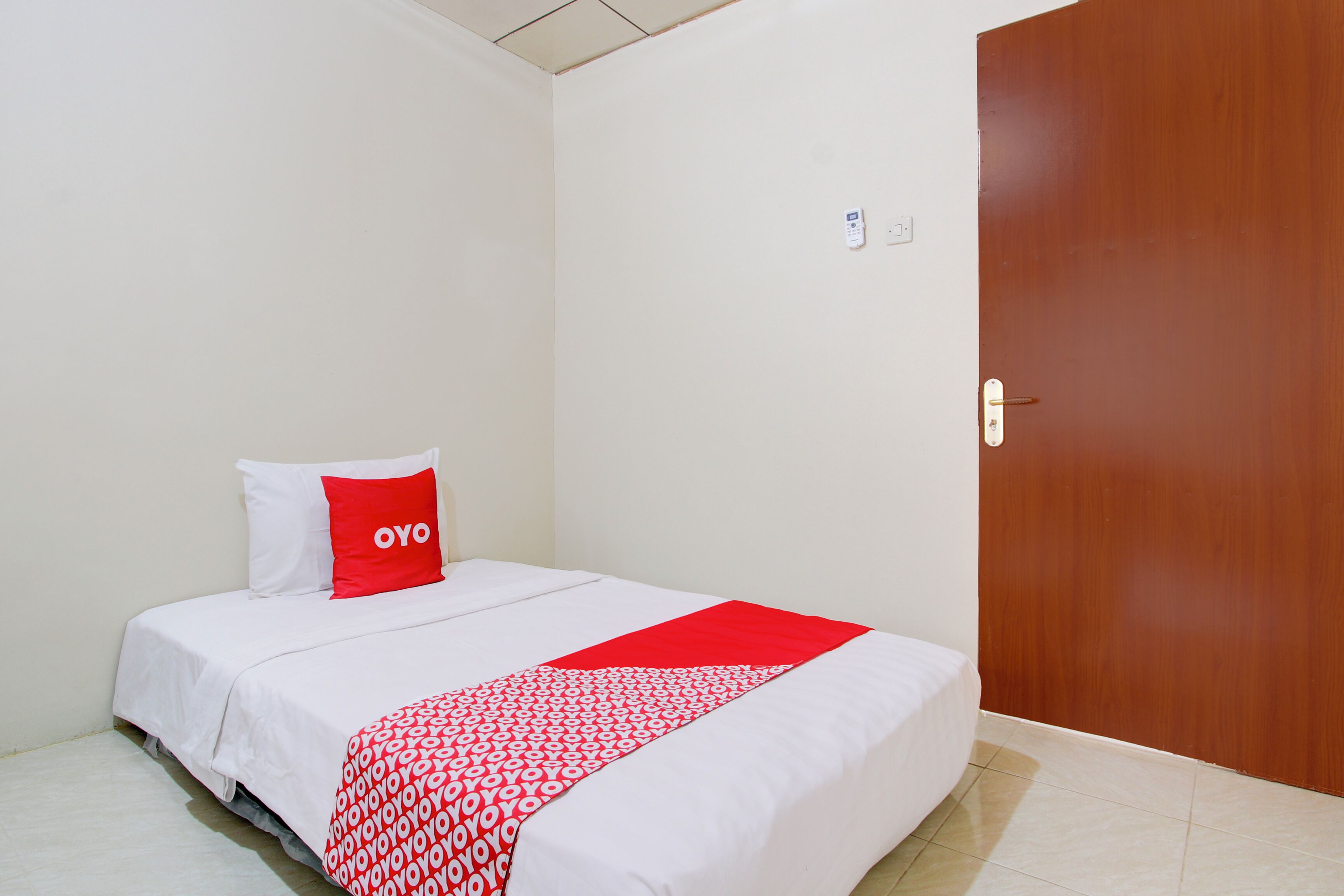Una cama bien hecha con una colcha blanca y roja, un almohadón rojo con el texto 'OYO' y una puerta de madera a la derecha.