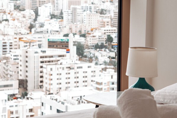 A hotel room with a view of a cityscape through a large window, a teal bedside lamp, and a neatly made bed.