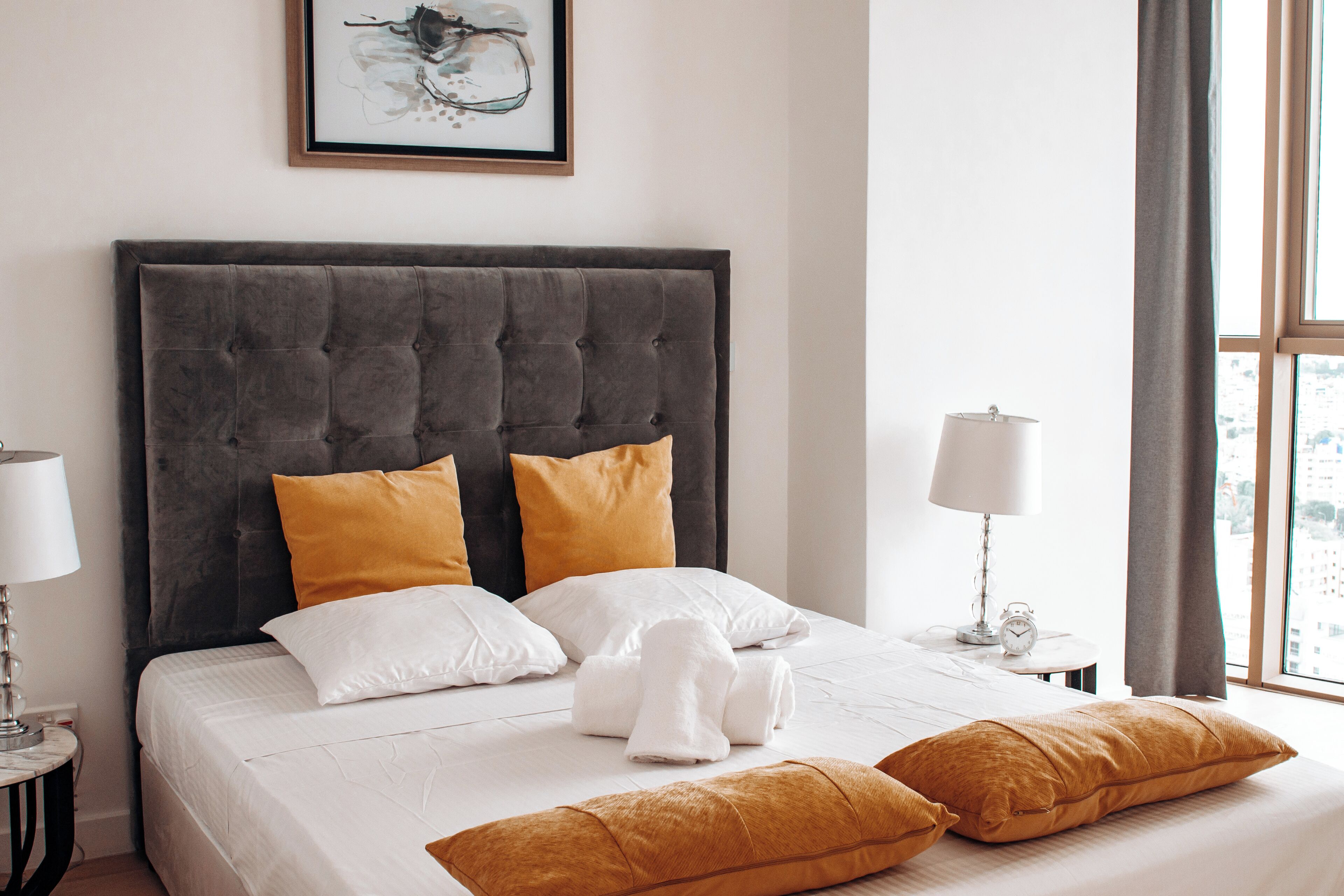 Una cama bien hecha con almohadones blancos y mostaza, un cabecero tapizado en gris y una obra de arte enmarcada encima.