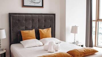 Una cama bien hecha con almohadones blancos y mostaza, un cabecero tapizado en gris y una obra de arte enmarcada encima.