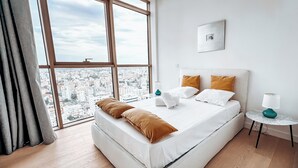Ein modernes Hotelzimmer mit großem Fenster, einem ordentlich bezogenen Bett, einem kleinen Nachttisch und Blick auf die Stadt.
