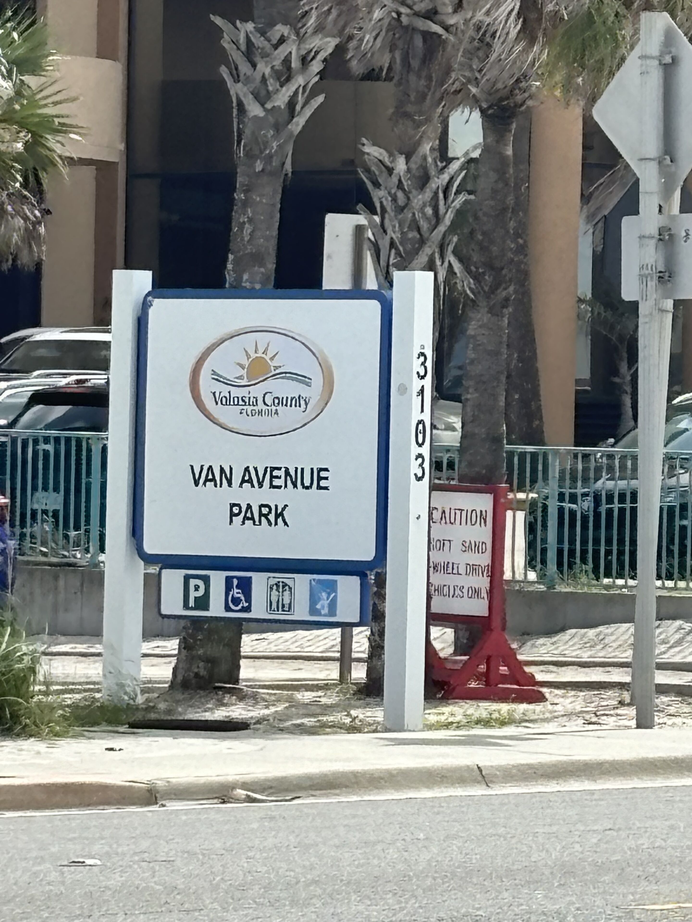 A sign for Van Avenue Park in Valusia County, Florida, with a wheelchair accessibility symbol and a caution sign nearby.