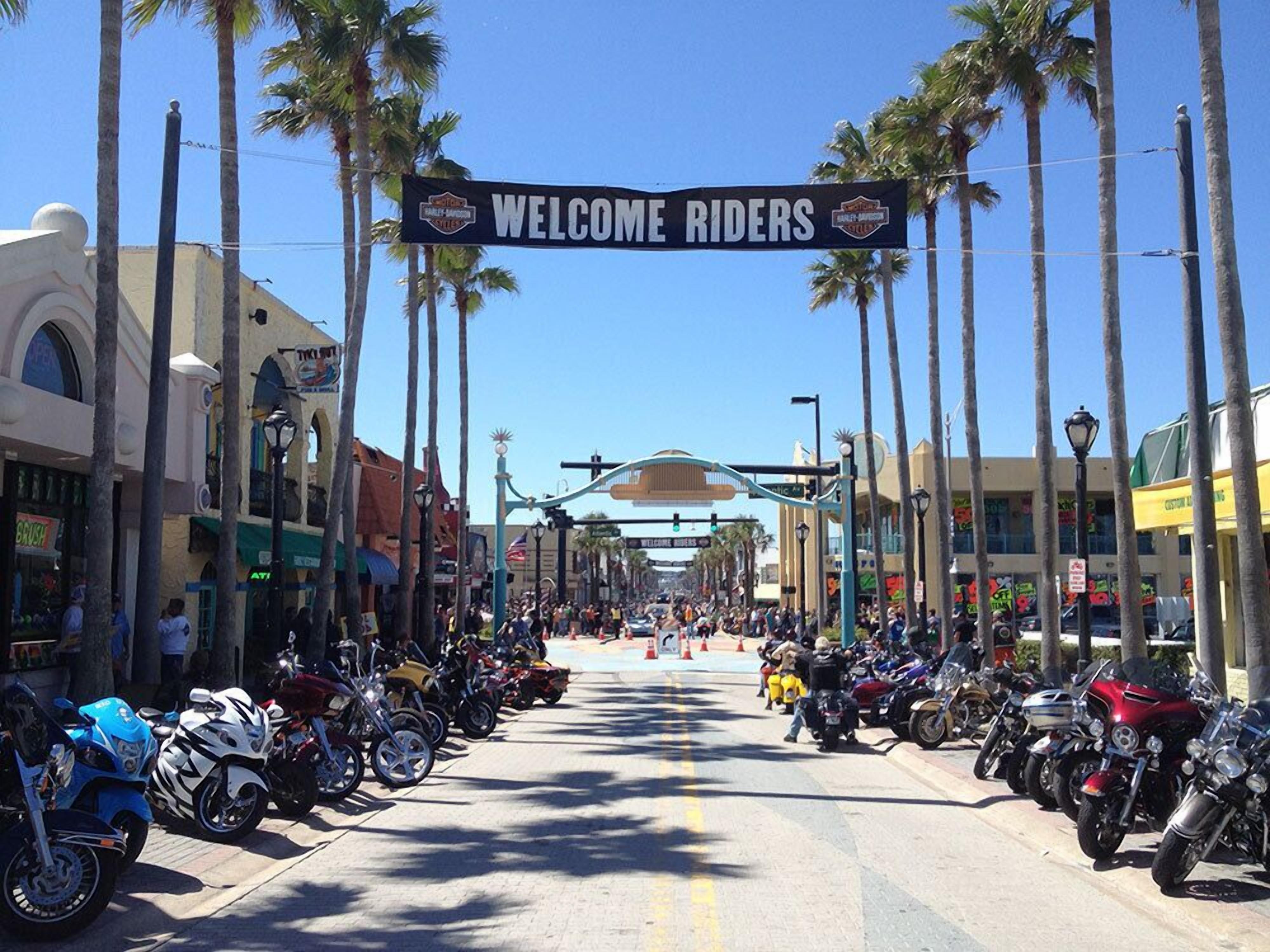 Una calle flanqueada por motocicletas y palmeras, con un cartel que dice 'Welcome Riders'.