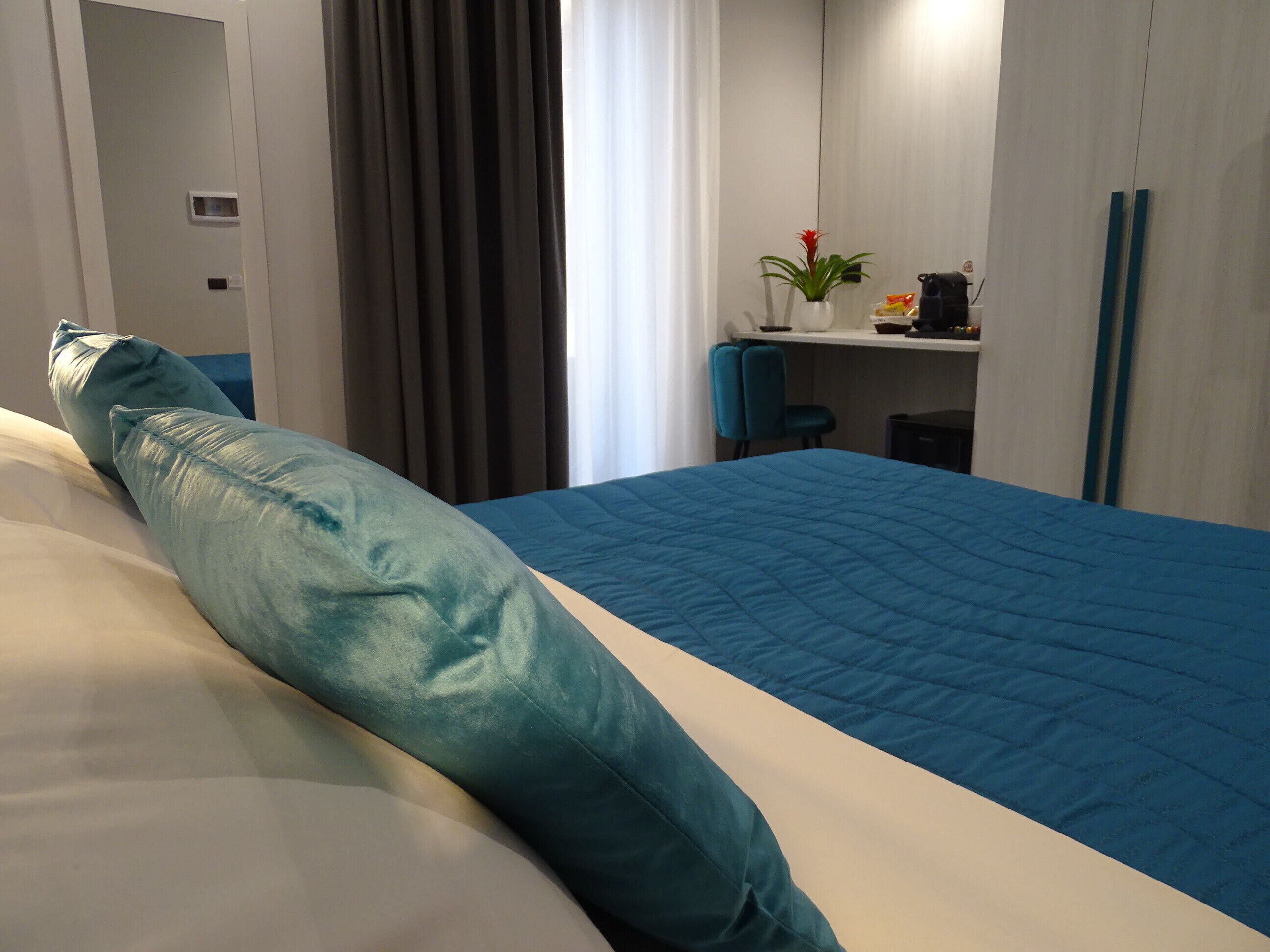A hotel room with a bed, a desk, a chair, a plant, and a shelf with items.