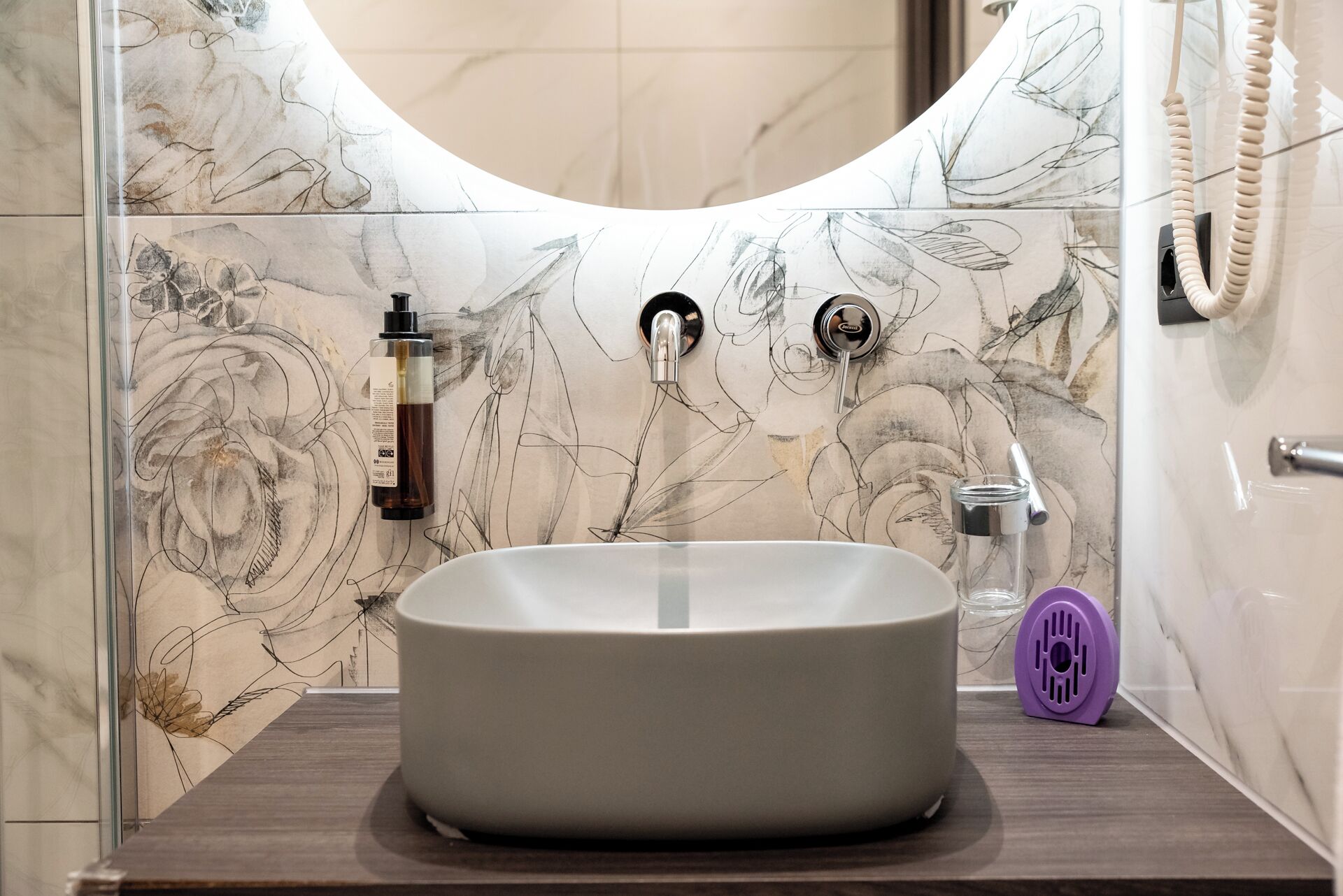A bathroom with a large oval sink, a handheld showerhead, and a glass bottle of liquid soap.