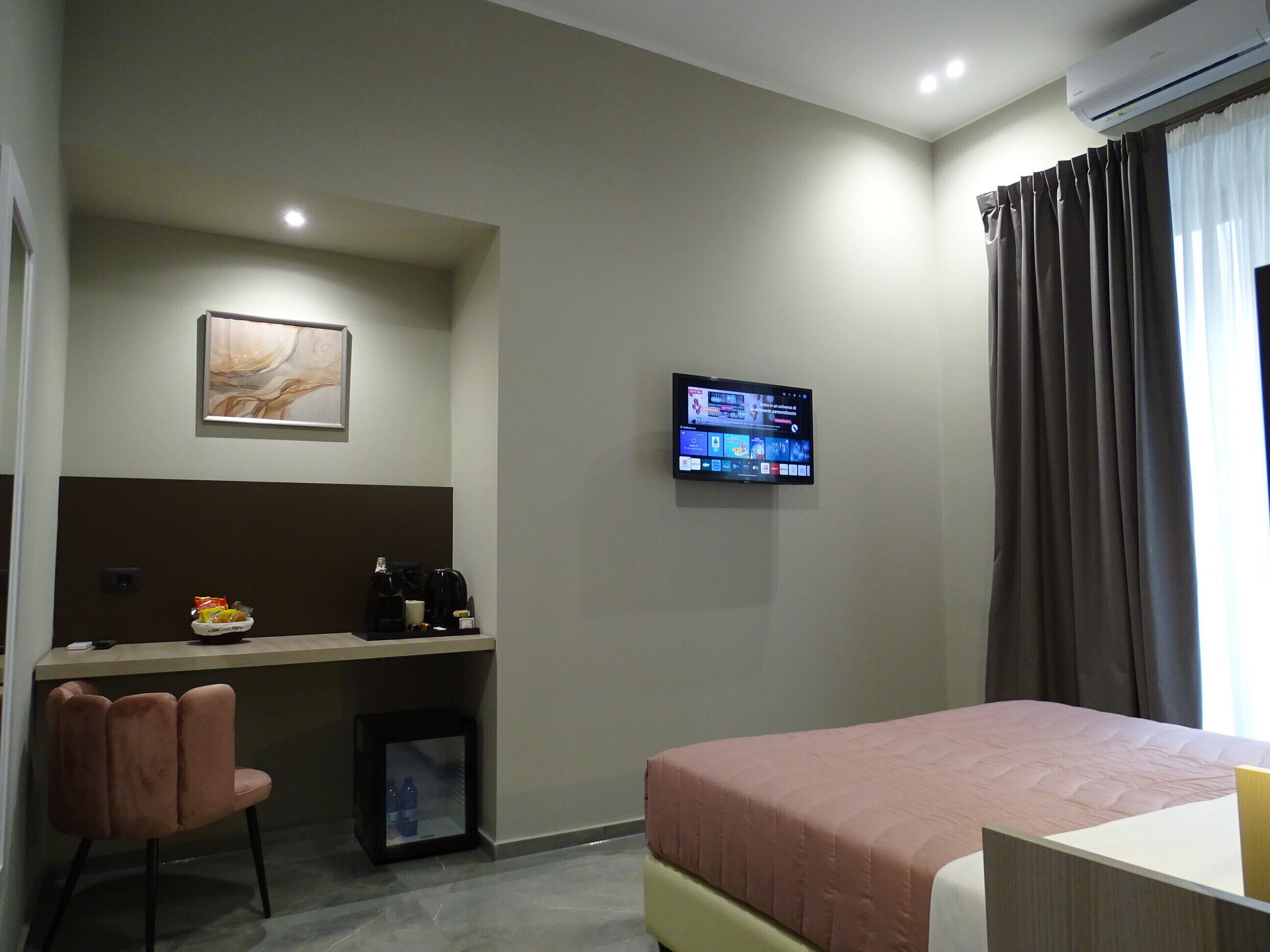 Una camera d'albergo con un letto, una scrivania con una sedia, una televisione a muro e un piccolo armadietto.