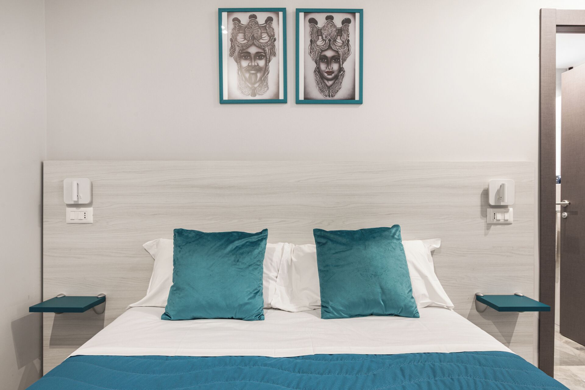 A modern hotel room with a large bed, teal and white bedding, two wall-mounted light switches, and two framed artworks above the bed.
