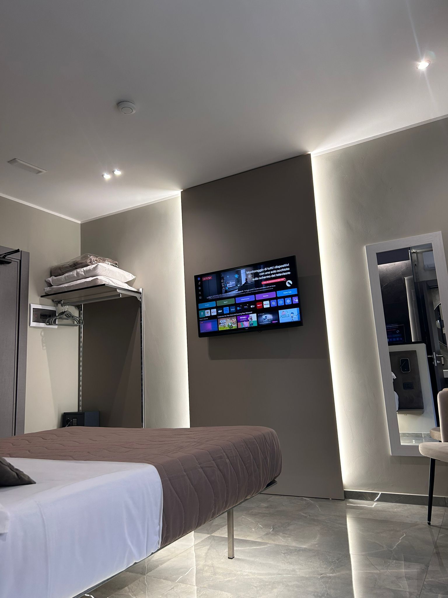 A modern hotel room with a bed, a flat-screen TV mounted on the wall, a wardrobe, and a small seating area.