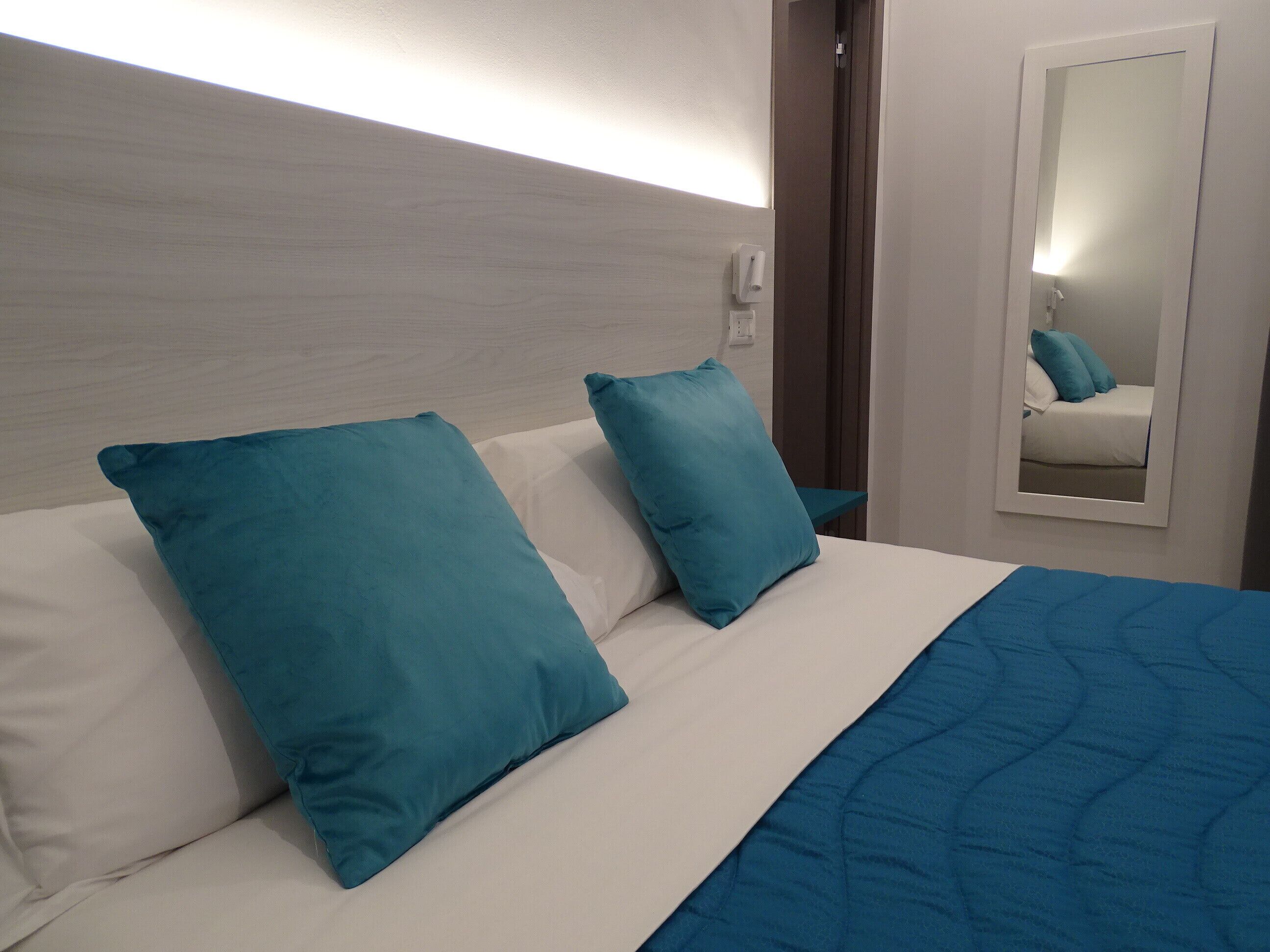 A hotel room with a bed, two teal pillows, a white duvet, and a blue blanket.