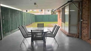 Un patio couvert avec une table et quatre chaises, une clôture verte et un mur en briques doté d’une porte-fenêtre coulissante.