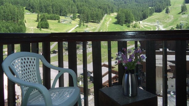 A balcony with a white chair and a vase of flowers, overlooking a mountainous landscape with green fields and a clear sky.