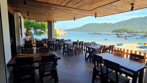 Un espace extérieur aménagé avec des tables et des chaises en bois, offrant une vue sur une plage où se trouvent des bateaux, le tout sur fond de montagnes.