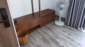 Une pièce avec un banc en bois, un ventilateur sur pied, un panier en osier et une porte dotée d’une poignée noire.