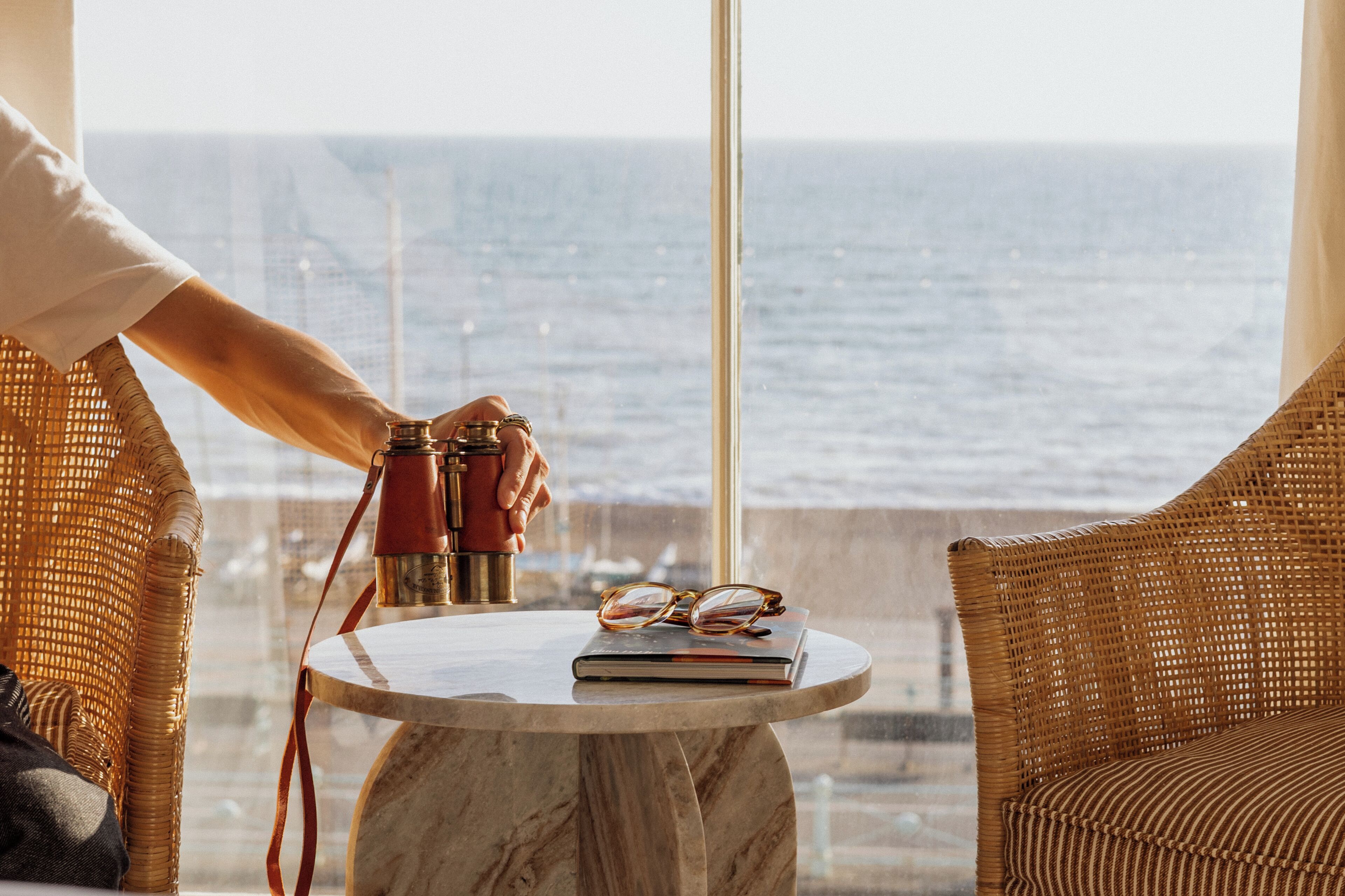 Une pièce avec vue sur l’océan, une table ronde sur laquelle reposent une paire de lunettes et un livre, et un fauteuil en osier.