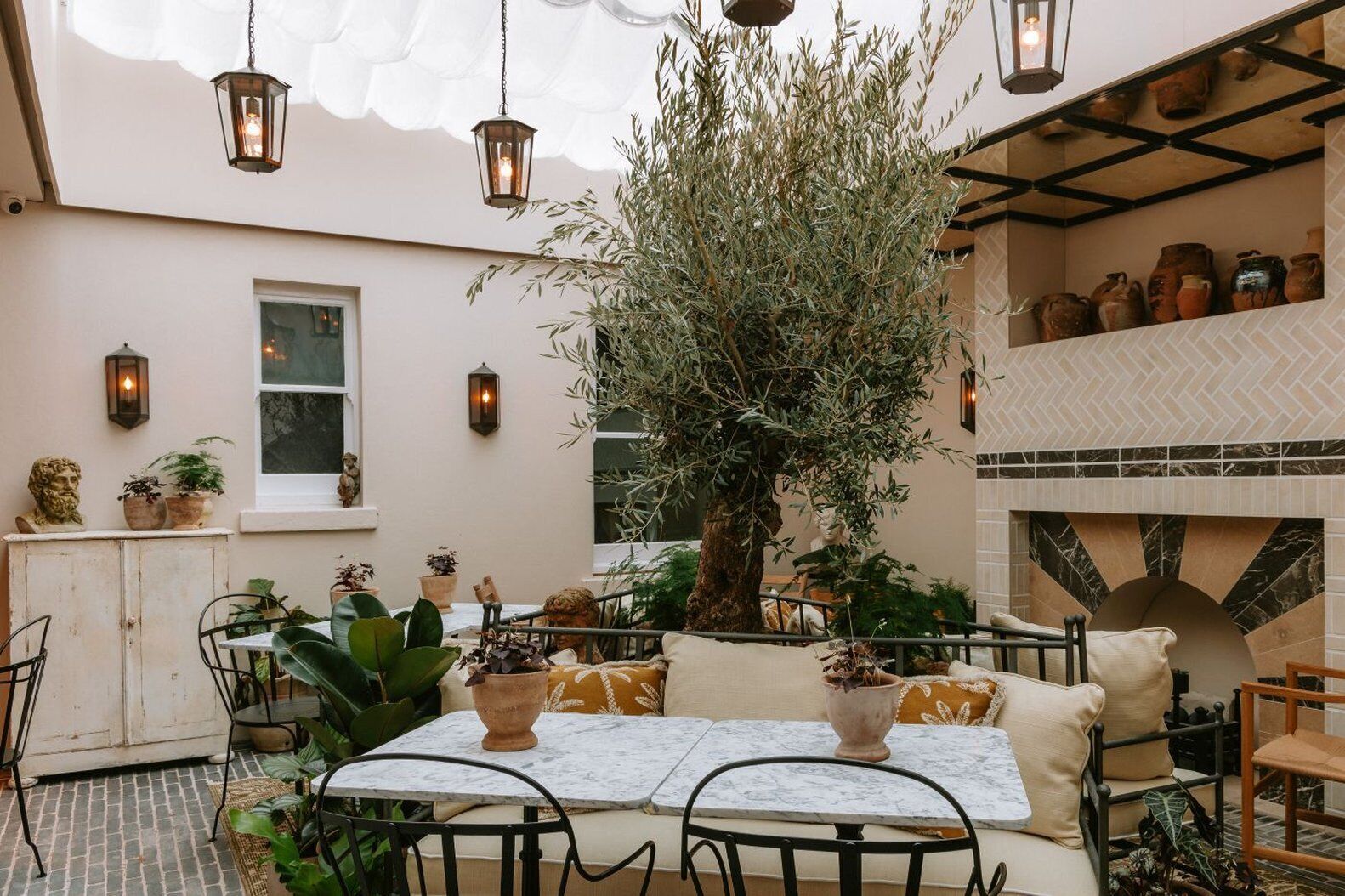 An outdoor patio with a table and chairs, surrounded by potted plants and a large tree.