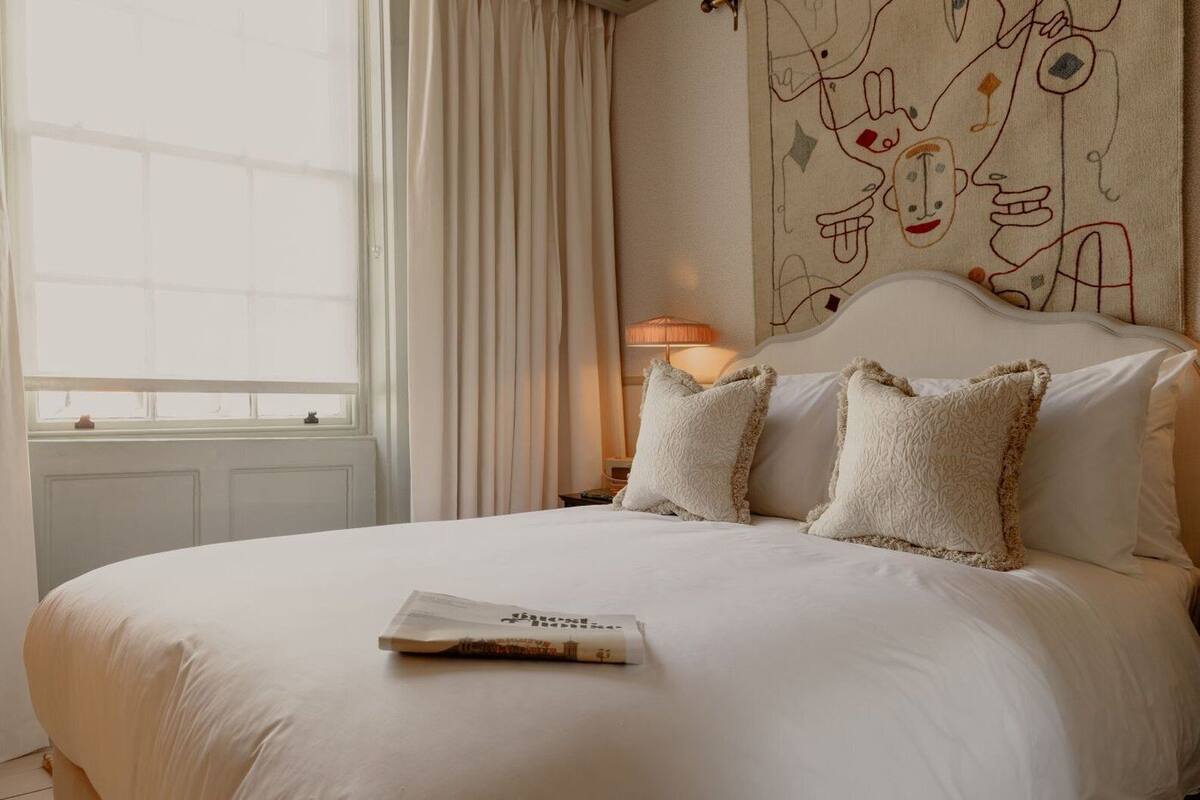 Una cama bien hecha con un almohadón y una revista, una lámpara y una ventana con cortinas.