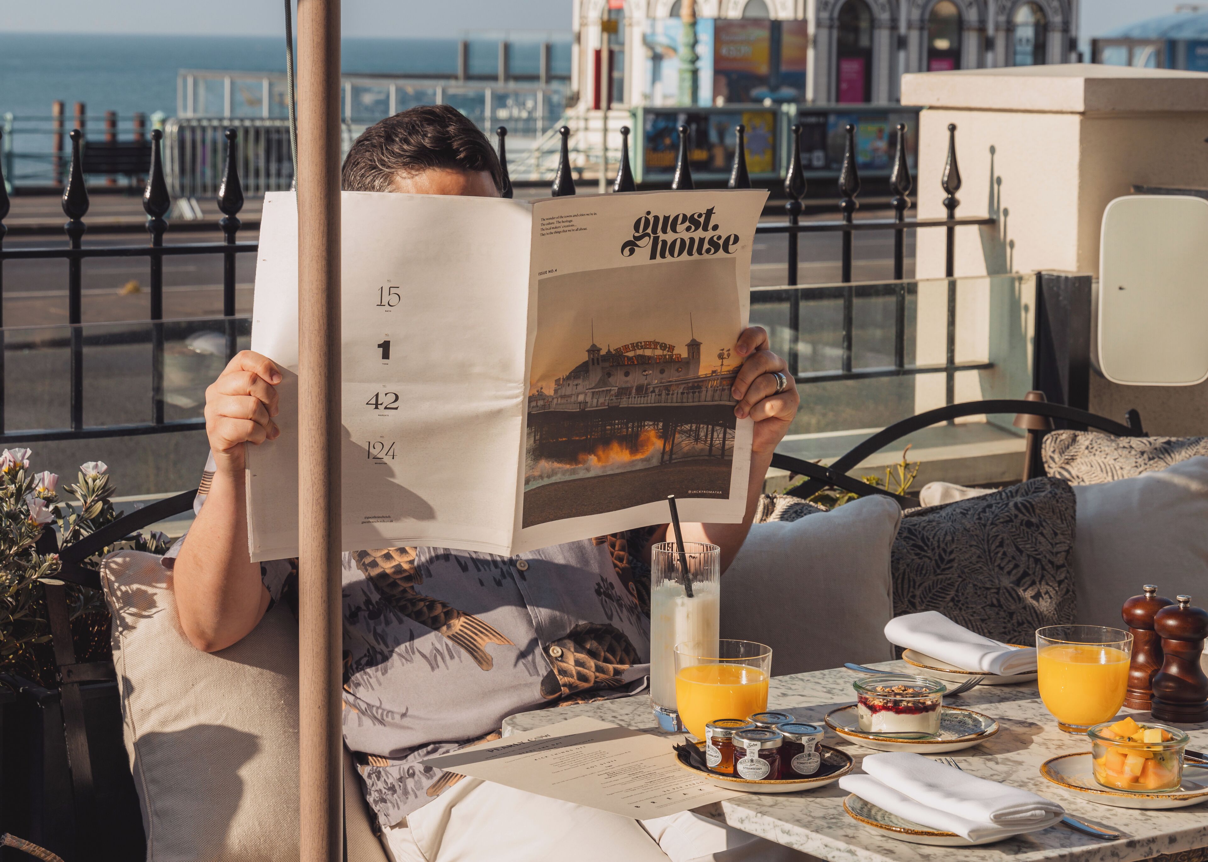 Une personne est assise à une table, devant un magazine, et profite de la vue sur la mer. Un bâtiment se trouve en arrière-plan.