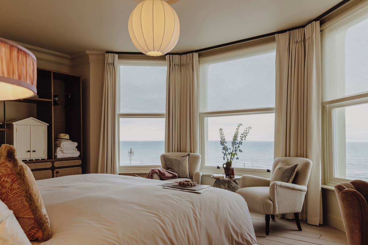 Un cuarto de hotel con una cama grande, dos sillones, una mesita de noche con un florero y vista al mar.