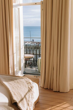 Une chambre avec un grand lit, une vue sur la mer à travers des rideaux ouverts, et un balcon avec une chaise et une balustrade.