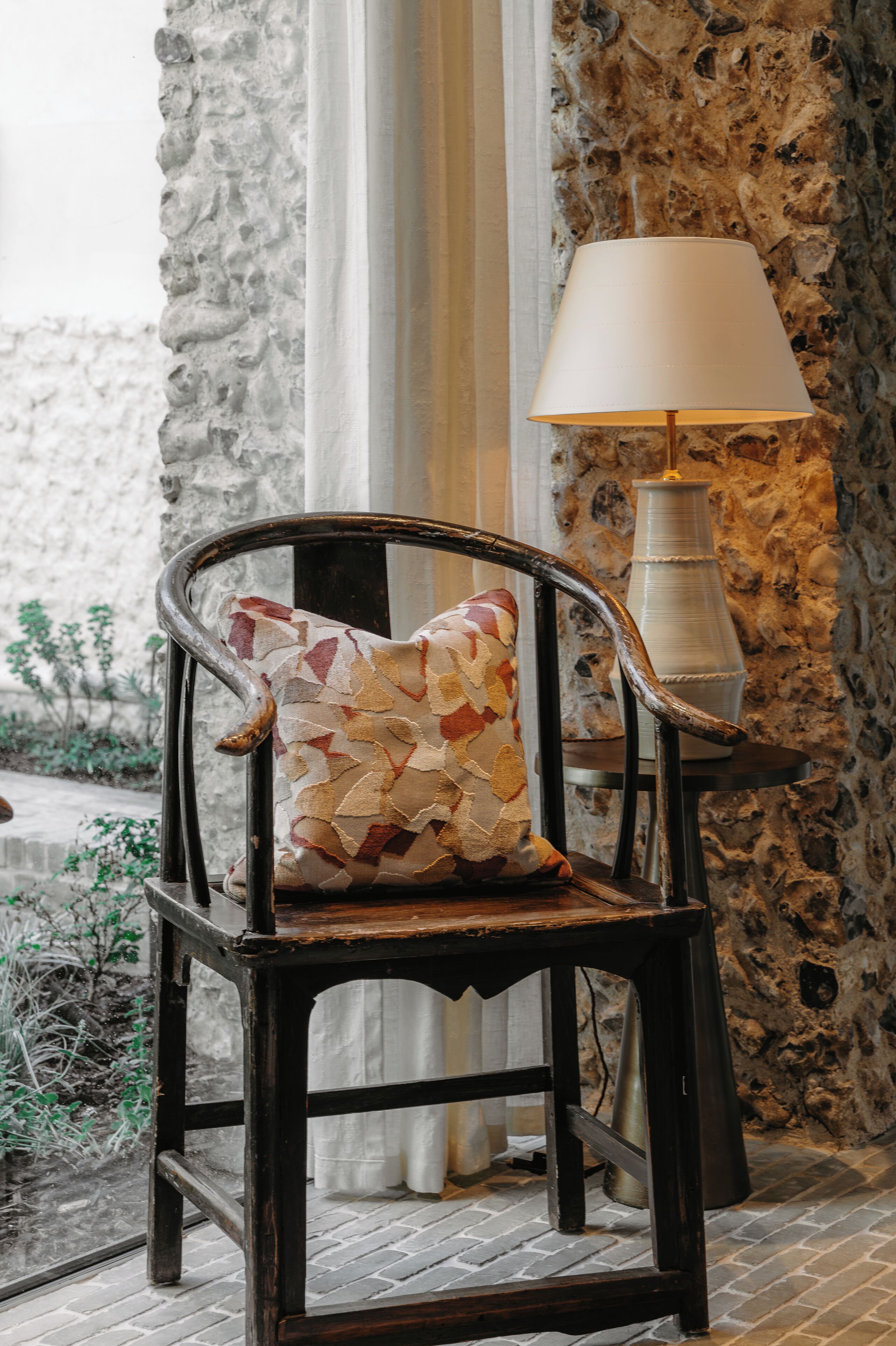 A rustic wooden chair with a patterned cushion, a table lamp, and a stone wall in the background.