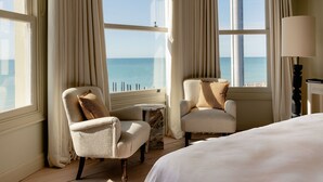 A hotel room with a bed, two armchairs, a small table, and a lamp. The room has large windows with curtains, offering a view of the sea. - No. 124 Brighton (Brighton)
