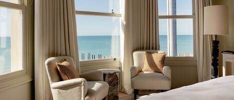 Quarto de hotel com cama, duas poltronas, uma mesinha e um abajur. O quarto possui grandes janelas com cortinas, proporcionando vista para o mar.