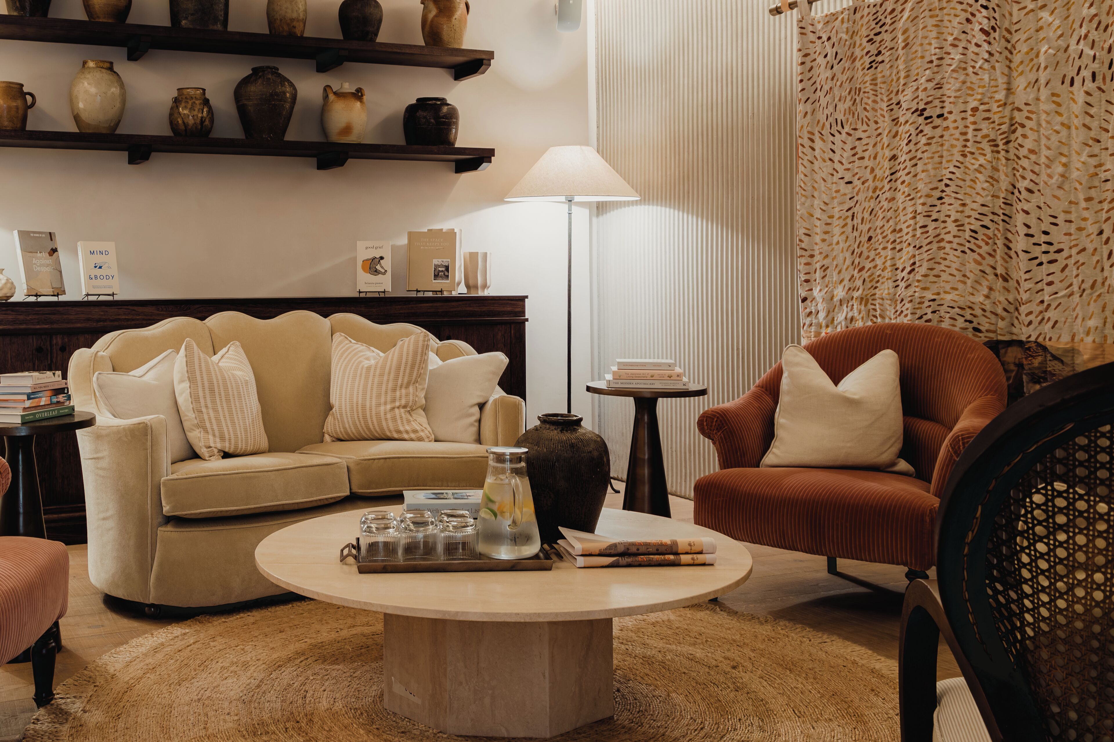 A cozy living room with a beige sofa, a brown armchair, a coffee table, and a bookshelf with decorative items.