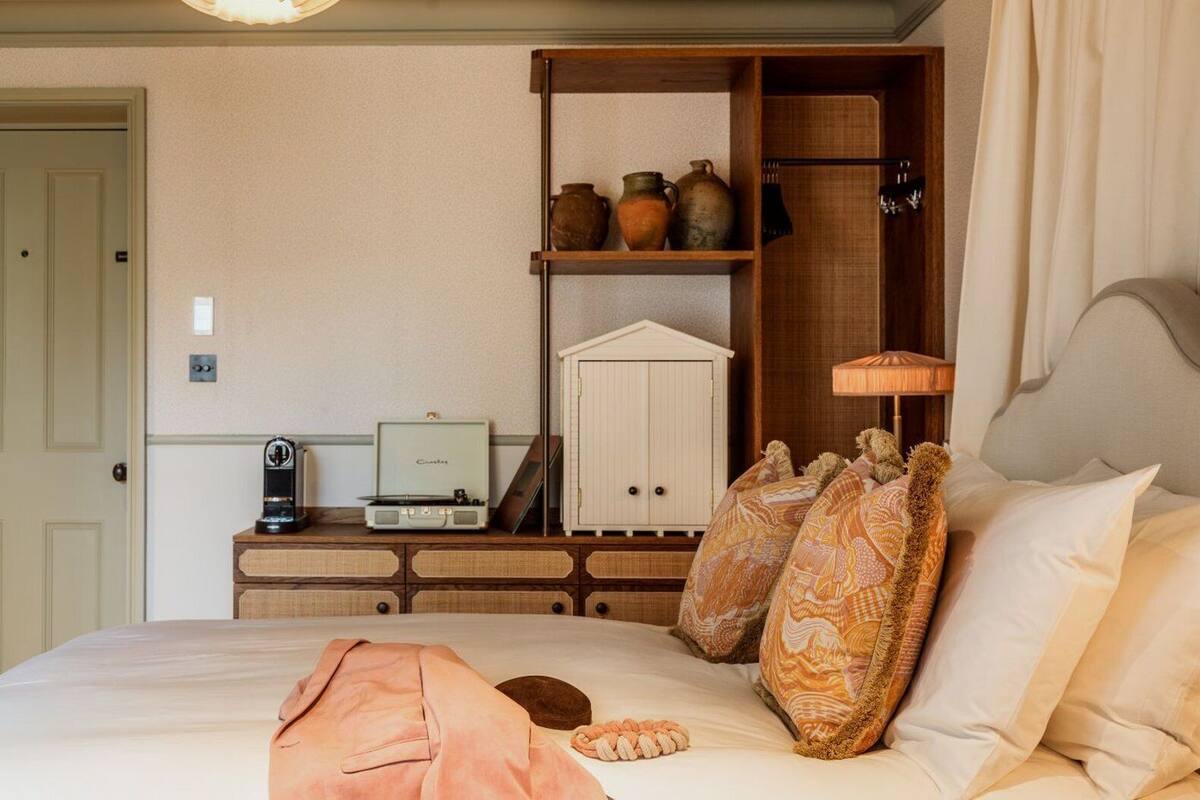 Una cama bien hecha con almohadones decorativos, una mesita de noche con una radio antigua y un aparador de madera con cerámica.