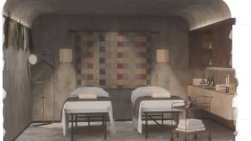 Two single beds with white linens sit against a textured wall, complemented by a colorful quilt and a small kitchenette with a wooden countertop.