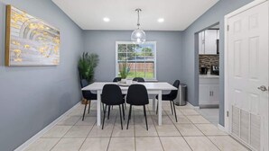Un espace repas avec une table blanche, des chaises noires, un luminaire suspendu, une fenêtre avec des stores et une porte donnant sur la cuisine.