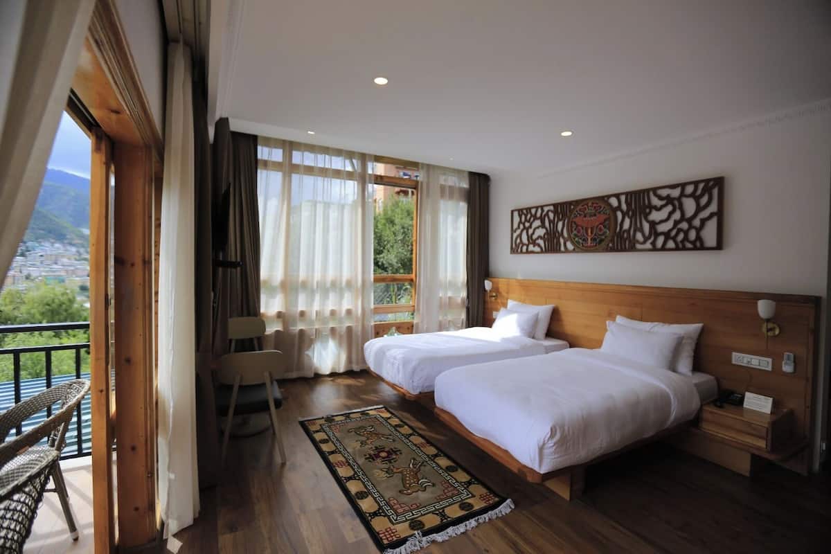 Une chambre d’hôtel avec deux lits, une tête de lit en bois, un élément décoratif mural, une vue sur la verdure par la fenêtre et un balcon avec des meubles en osier.