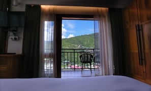 Habitación de hotel con balcón, cama, silla y vista a una colina verde con un edificio en la cima.