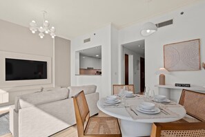 Un salon moderne doté d’un téléviseur à écran plat, d’un canapé, d’une table à manger pour quatre personnes et d’un coin cuisine visible à travers une porte ouverte.
