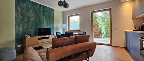 Una sala moderna con sofá, televisor, mesita de madera y escritorio. Hay una ventana con vista a árboles y una puerta que da a un balcón.