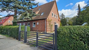A red brick house with a sloped roof and multiple windows, surrounded by a hedge and a metal gate. - Quiet 2 room apartment with WLAN on the first floor - MH5 (Markgrafenheide)