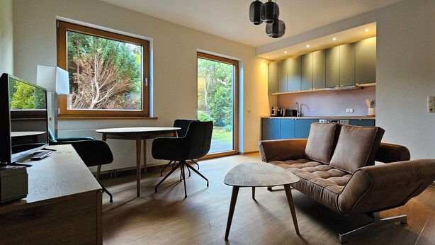 A modern living room with a sofa, a dining table, and a TV. There is a kitchen area with cabinets and a window. - Quiet 2 room apartment with WLAN on the first floor - MH5 (Markgrafenheide)