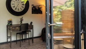 A room with a wall clock, a desk with a chair, a small table with a vase, and a door leading to an outdoor area with a table and chairs.