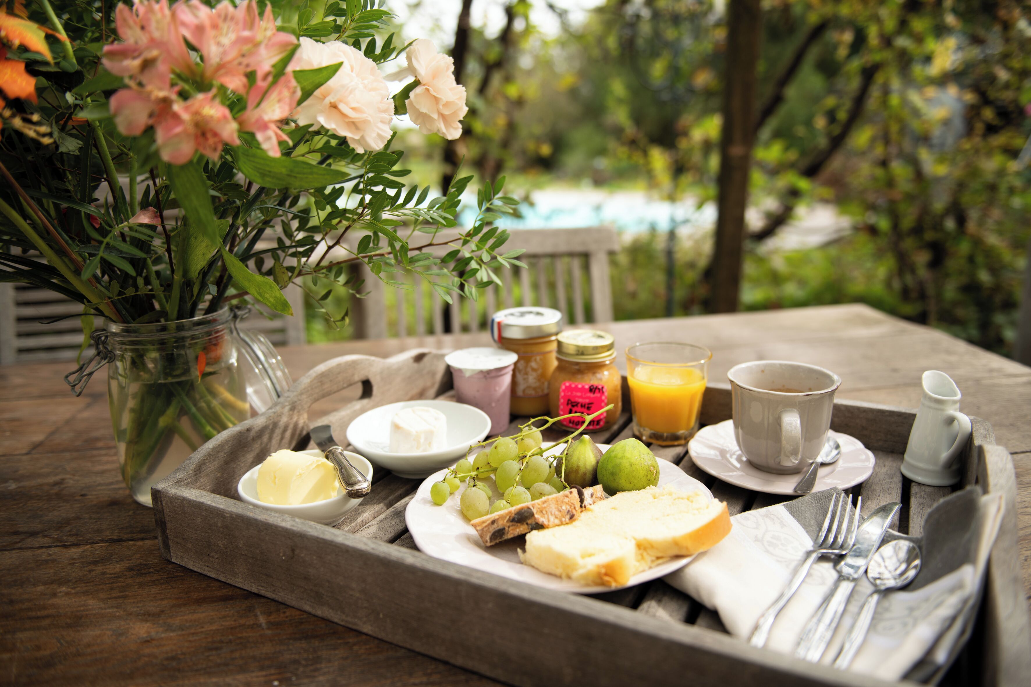 Una bandeja de madera con productos de desayuno como queso, uvas y mantequilla, junto a un florero con flores, sobre una mesa de madera al aire libre.
