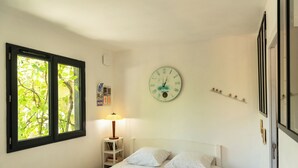 A bedroom with a bed, a wall clock, a window with a view of trees, a bedside lamp, and a small shelf with books.
