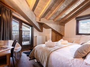 Une chambre avec un plafond en bois, un grand lit recouvert de draps blancs, une chaise, une table et une vue sur l’extérieur par une fenêtre.