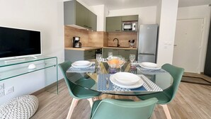 Une cuisine compacte dotée d’appareils électroménagers modernes, un coin repas avec une table en verre et des chaises vertes, et un téléviseur à écran plat posé sur une étagère en verre.