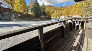 Une terrasse en bois avec une rambarde donnant sur une route où circule une voiture, le tout entouré d’arbres aux couleurs automnales et sous un ciel dégagé.