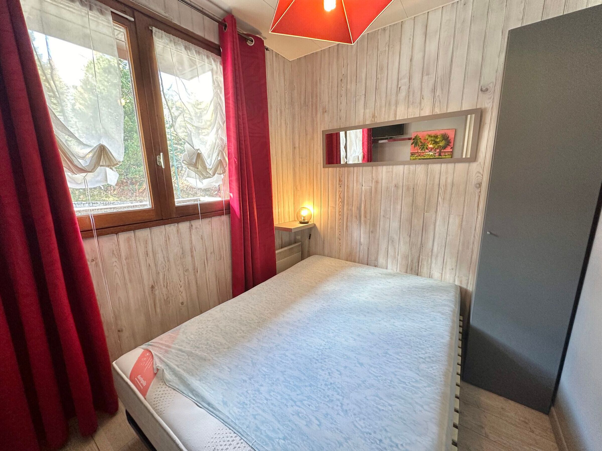 Une pièce avec un lit, une fenêtre avec des rideaux, une petite étagère avec une plante et un mur en bois.