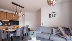 Ein modernes Wohnzimmer mit grauem Sofa, einem Essbereich mit grauen Stühlen und einer Küche mit Holzausbau und Pendelleuchten.