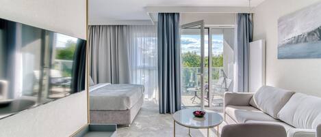 Una habitación de hotel moderna con cama, sofá, una mesita y televisor. Cuenta con un balcón con vistas al exterior.