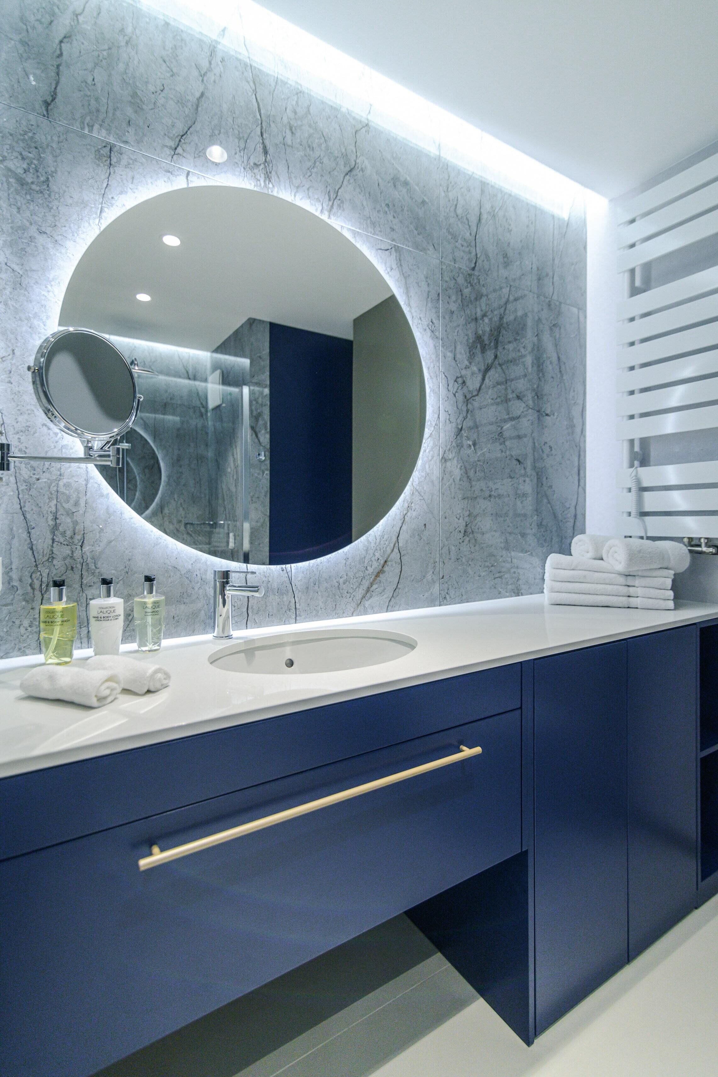 A modern bathroom with a marble wall, a round mirror, a white sink, and blue cabinets.